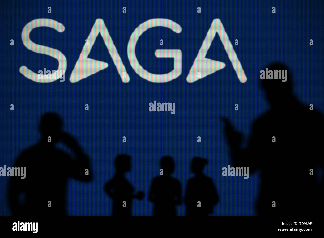 The Saga Insurance logo is seen on an LED screen in the background while a silhouetted person uses a smartphone in the foreground (Editorial use only) Stock Photo