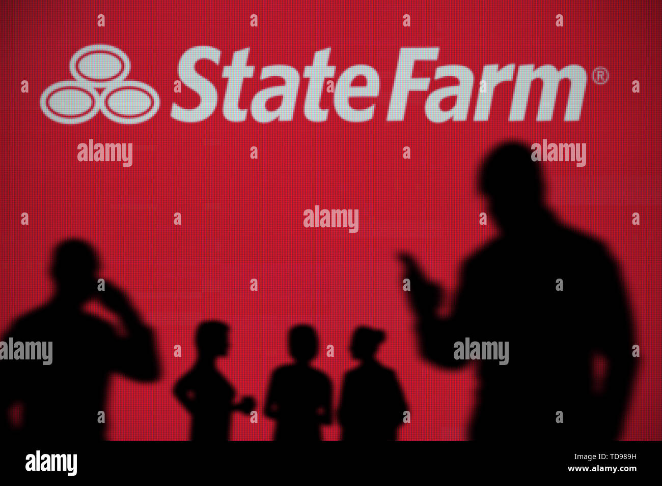 The State Farm logo is seen on an LED screen in the background while a silhouetted person uses a smartphone in the foreground (Editorial use only) Stock Photo
