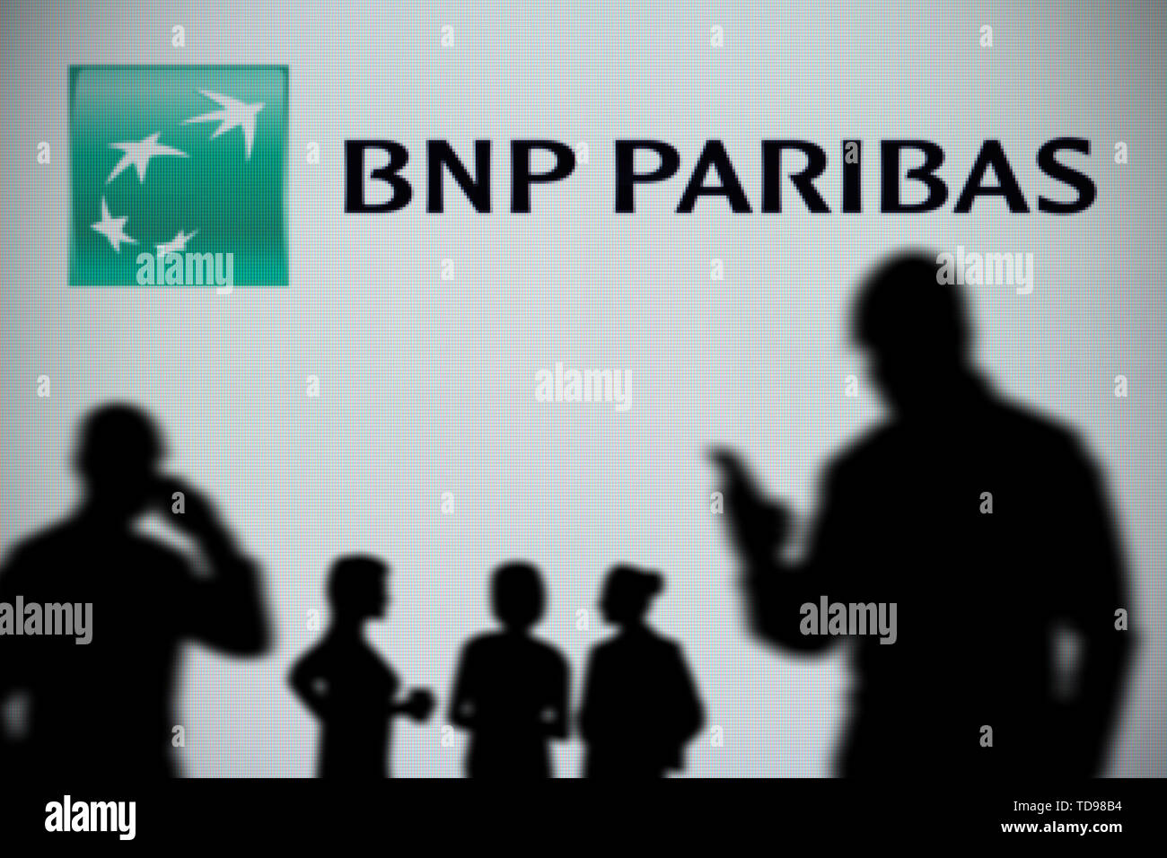 The BNP Paribas logo is seen on an LED screen in the background while a silhouetted person uses a smartphone in the foreground (Editorial use only) Stock Photo