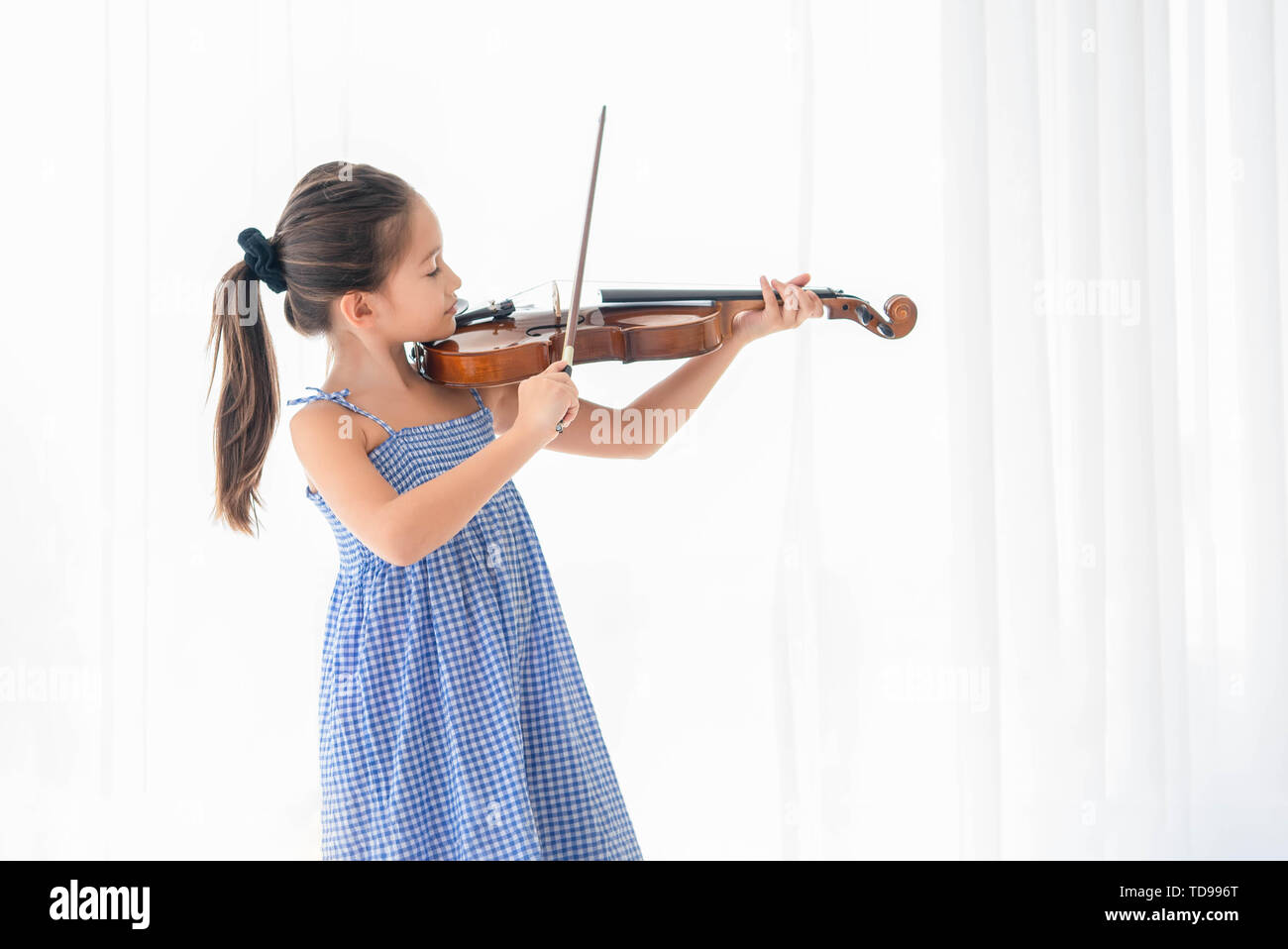 Cute girl playing violin in white bedroom with white curtain background. Musical and people lifestyles. Education and recreation concept. Back to scho Stock Photo