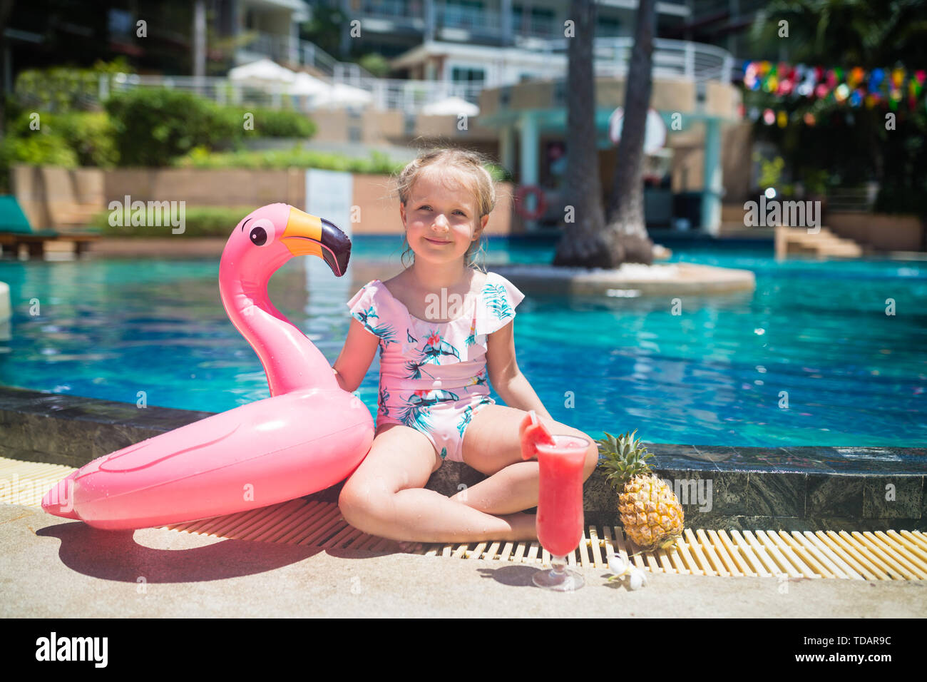 Cute little girl in swimsuit sitting near swimming pool with inflatable