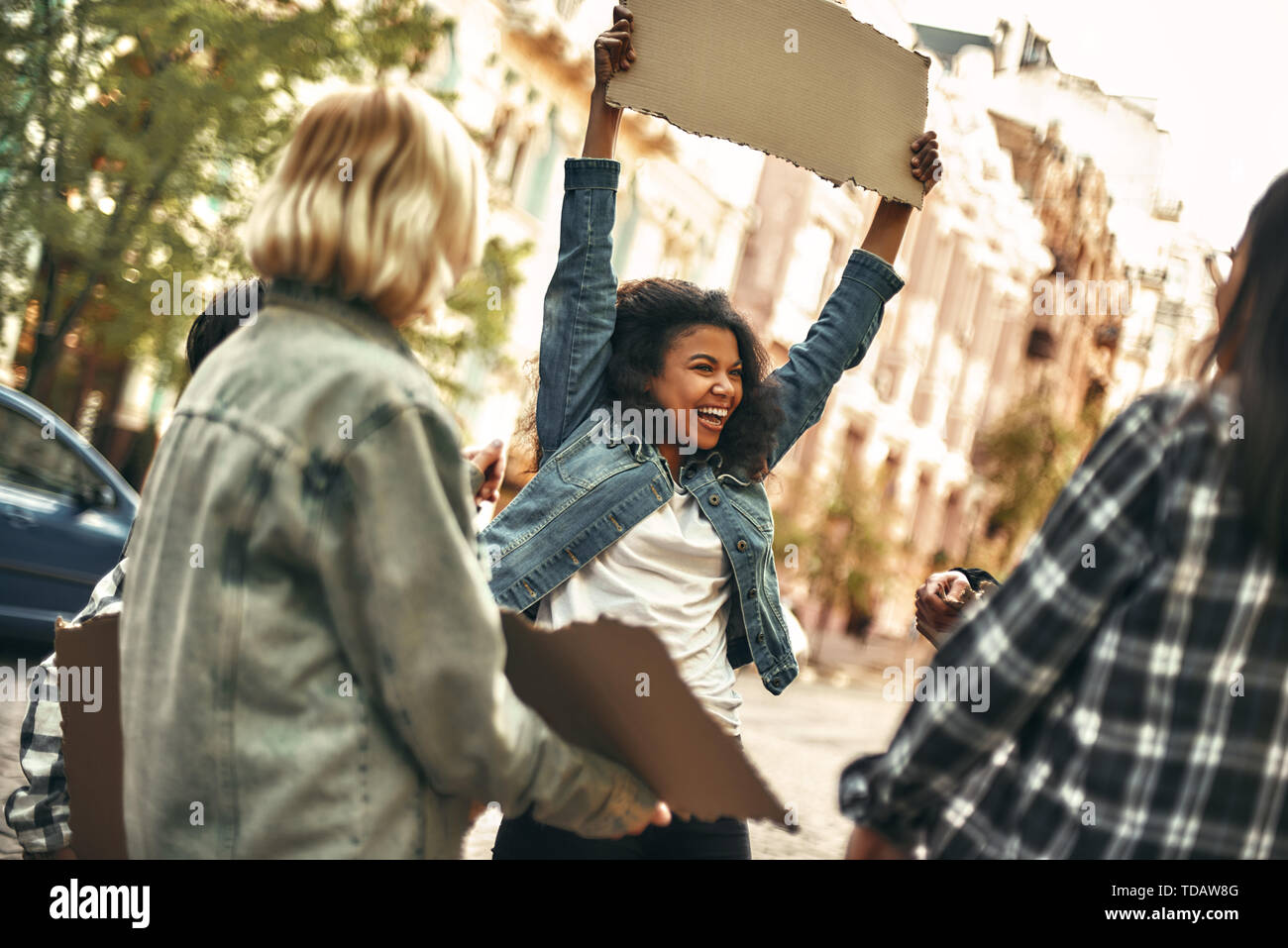 Feeling happy. Cheerful young afro american woman holding blank sign board over her head and laughing while standing on the road around other female a Stock Photo