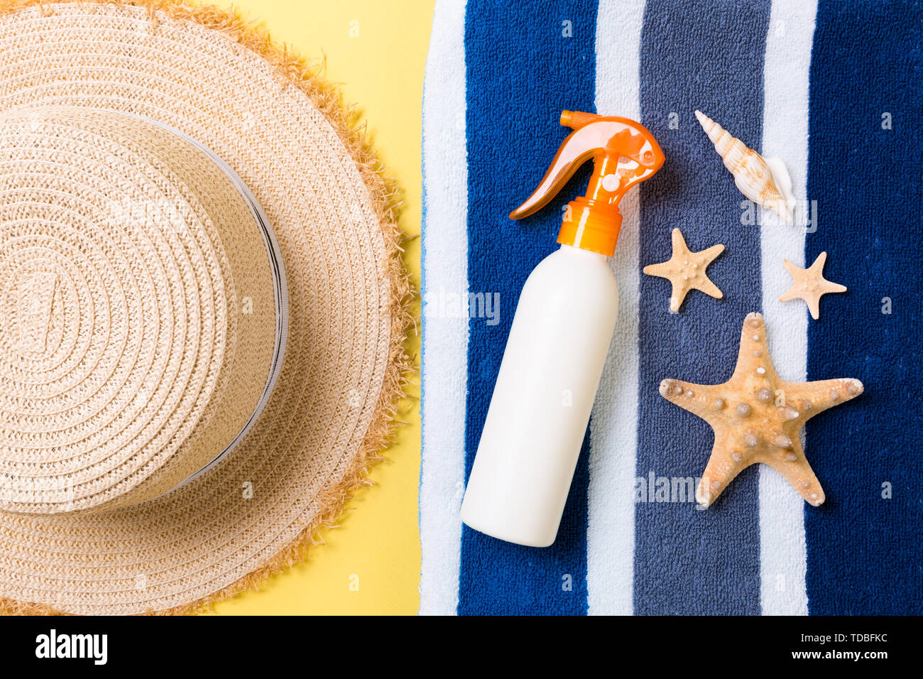 Beach flat lay accessories with copy space. Striped blue and white towel, seashells, staw sunhat and a bottle of sunblock on yellow background. Summer Stock Photo