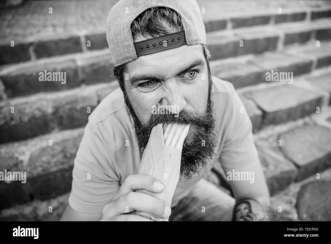 Junk food. Carefree hipster eat junk food while sit stairs. Guy eating hot dog. Snack for good mood. Unleashed appetite. Street food concept. Man bearded eat tasty sausage. Urban lifestyle nutrition. Stock Photo