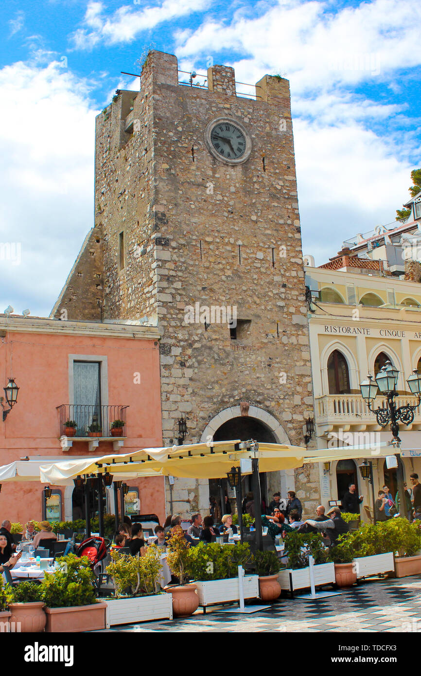 Taormina, Sicily, Italy - Apr 8th 2019: Tourists in restaurants and cafes gardens on historical Piazza IX Aprile square in front of famous Clock Tower. Sicilian city, tourist place, travel spot. Stock Photo