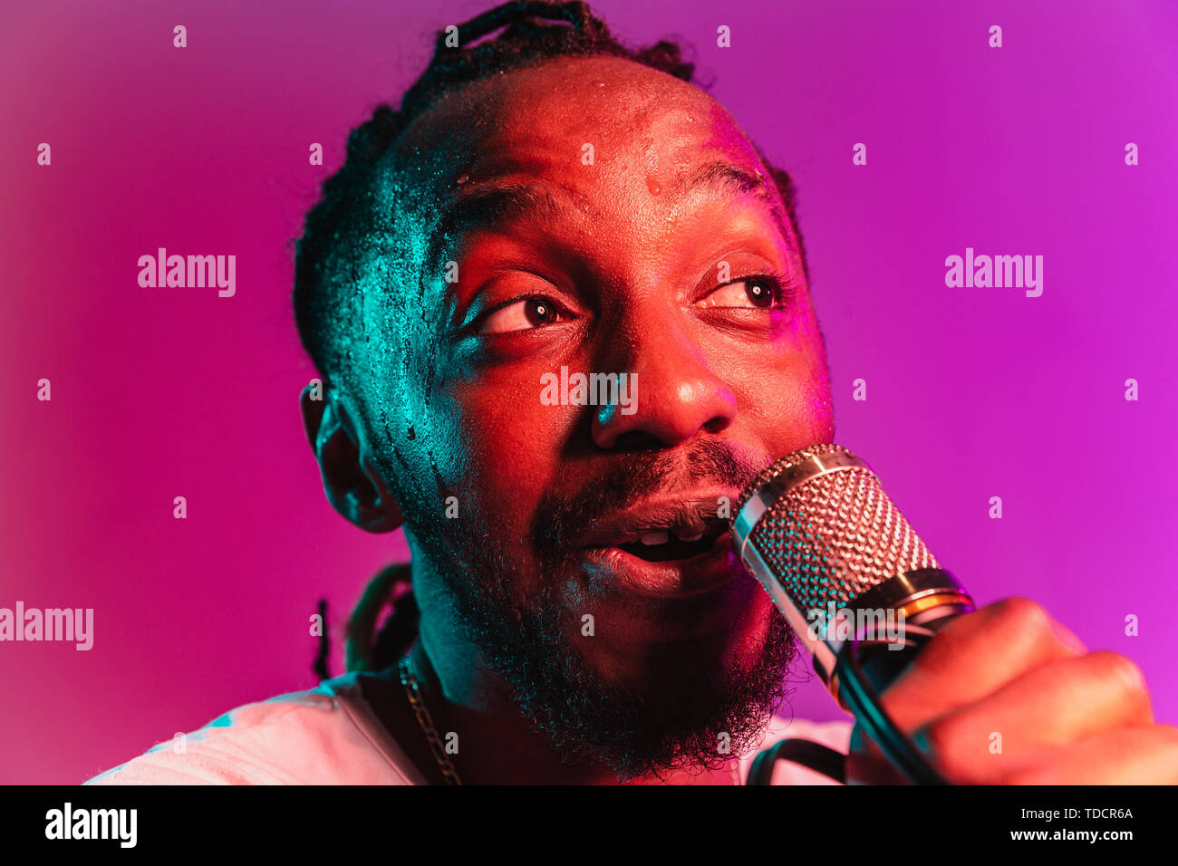 Young african-american jazz musician with microphone singing a song on purple studio background in trendy neon light. Concept of music, hobby, inspirness. Colorful portrait of joyful attractive artist. Stock Photo