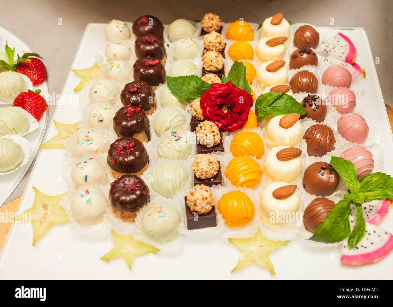 Small assorted cakes lined up decorated on dessert buffet with red rose and mint leaves, with slices of Carambola fruit Stock Photo