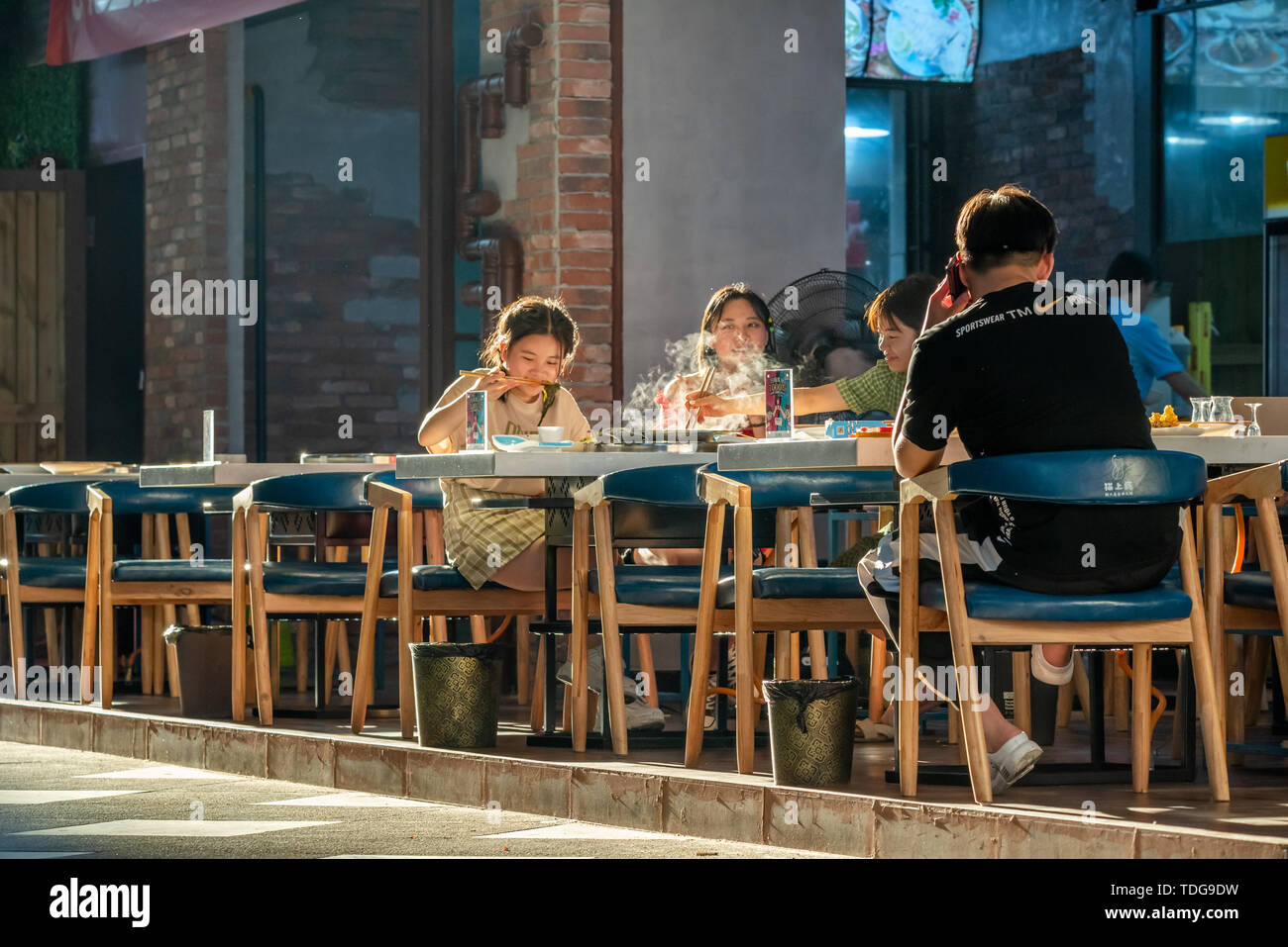 Chengdu, Sichuan province, China - June 12, 2019 : Young chinese people eating outside a restaurant in a street in late afternoon Stock Photo