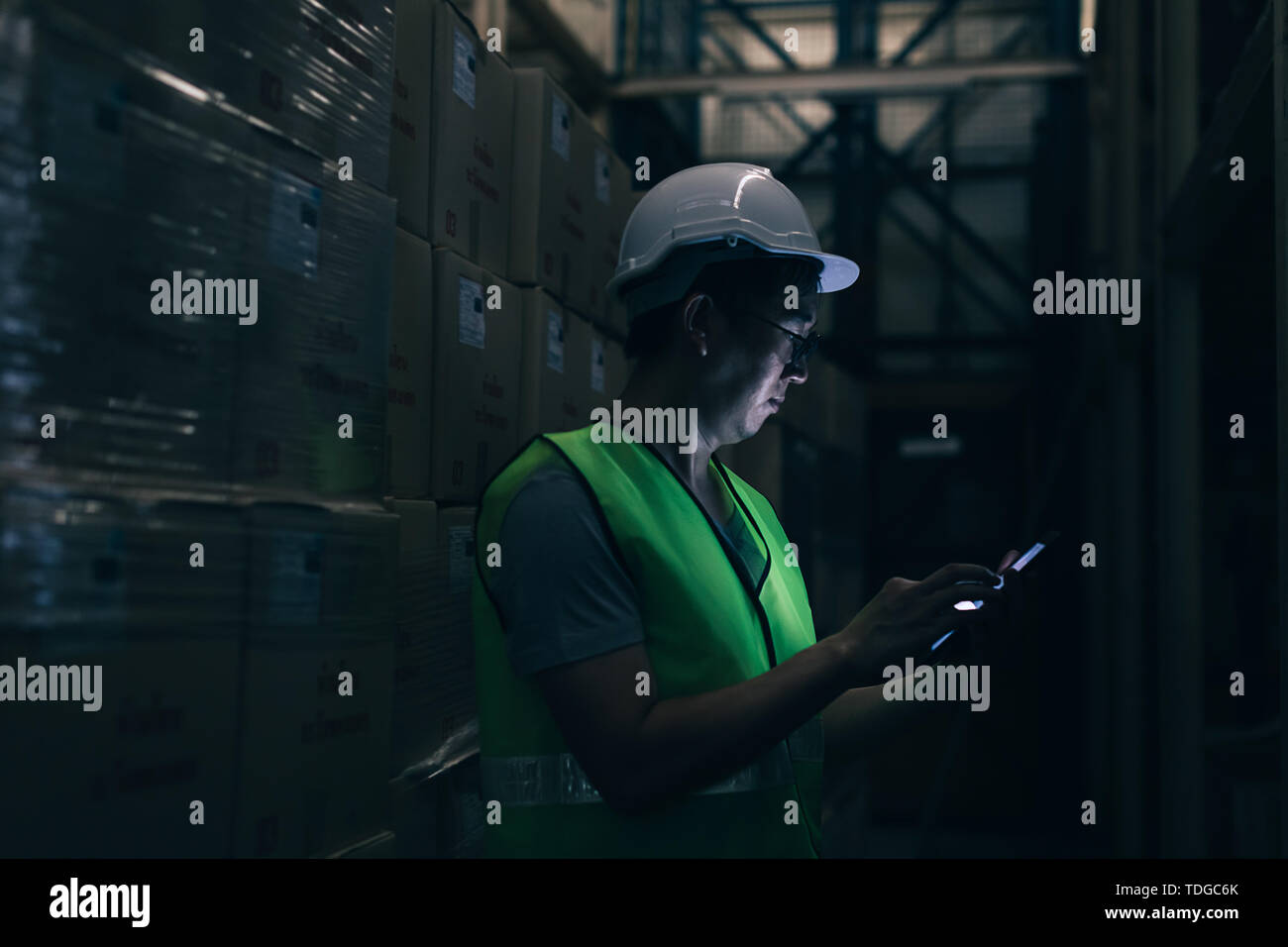 Side view of Asian young warehouse worker man with safety hard hat is checking order details with a digital tablet at inventory room Stock Photo