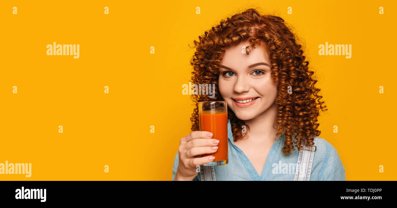 curly red-haired woman with a glass of fresh carrot juice on a yellow background. Try a healthy vitamin juice Stock Photo