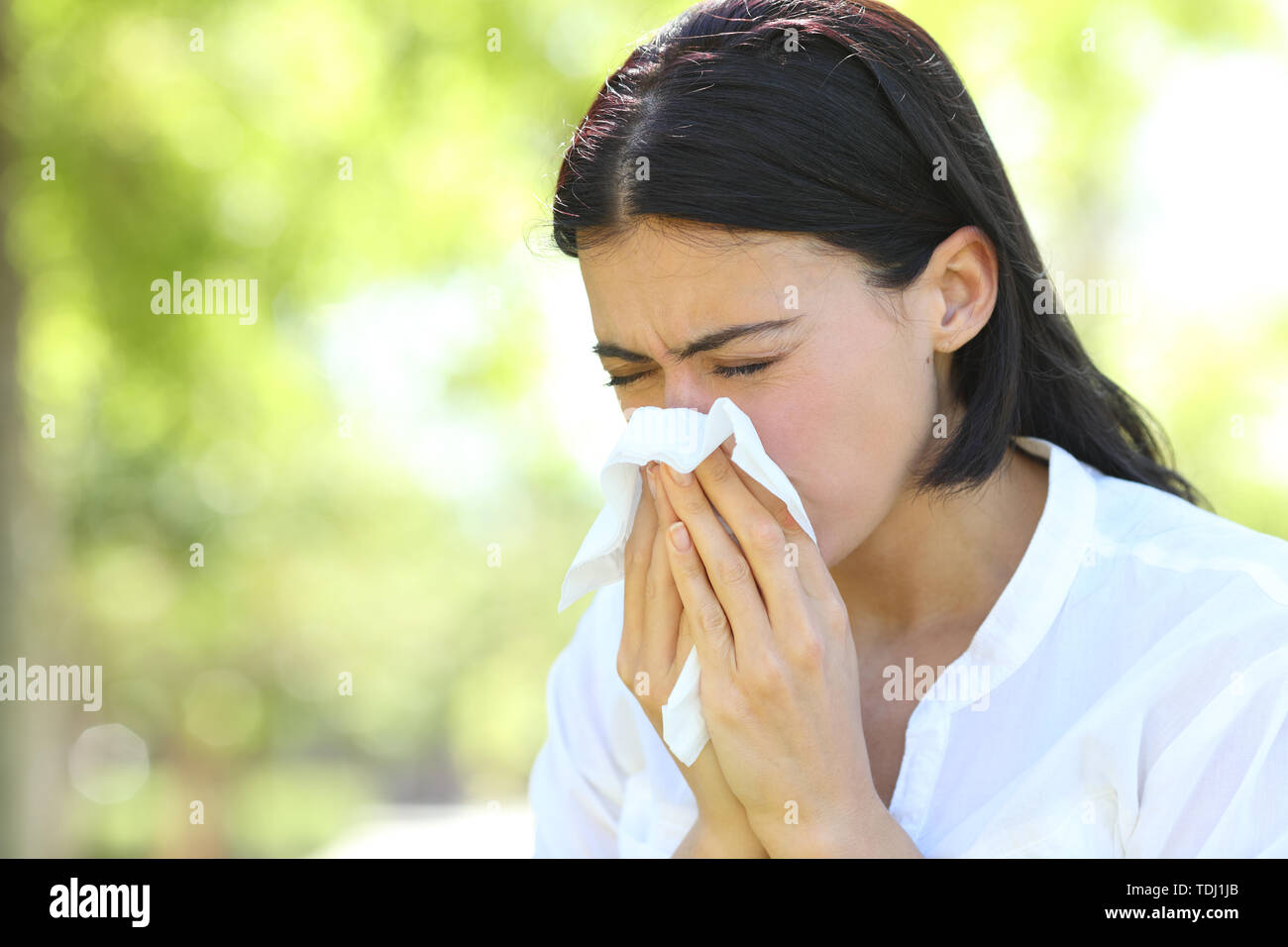 Ill woman sneezing covering mouth with a wipe standing in a park with a green background Stock Photo
