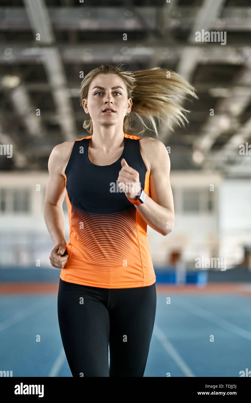 Incredible girl with flying hair in a black-orange T-shirt with dark pants  runs on the running track at the indoor stadium. She looks forward. Vertica  Stock Photo - Alamy