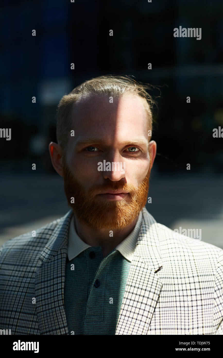 Portrait of serious confident businessman with red mustache and beard wearing fashionable checkered jacket standing in shade outdoors Stock Photo