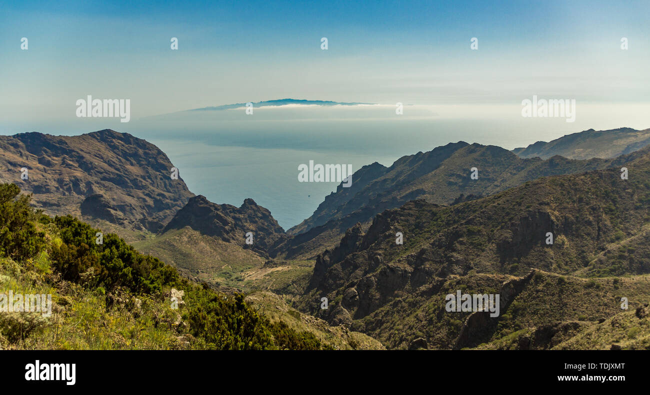 Island of La Gomera, soaring over the horizon, partly covered by the clouds. Bright blue sky. View from 1900 meters of altitude. Deep gorges Teno Natu Stock Photo