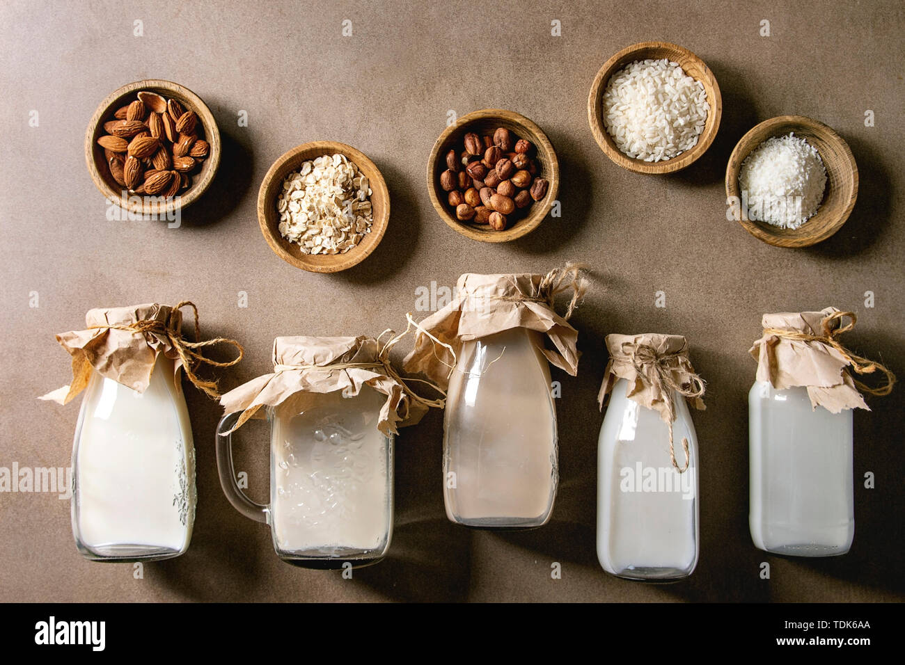 Variety of non-dairy vegan lactose free nuts and grain milk almond, hazelnut, coconut, rice, oat in glass bottles in row with ingredients above over b Stock Photo