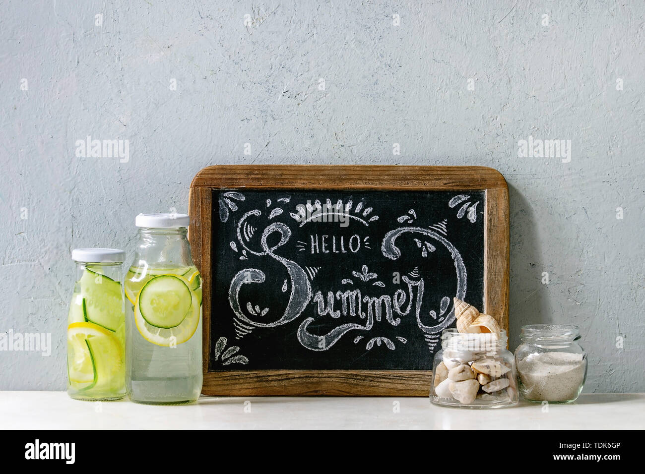 Summer theme. Chalk lettering Hello summer on vintage chalkboard, seashells and sand in glass jars, sunglasses, two glass bottles with sassy water on Stock Photo