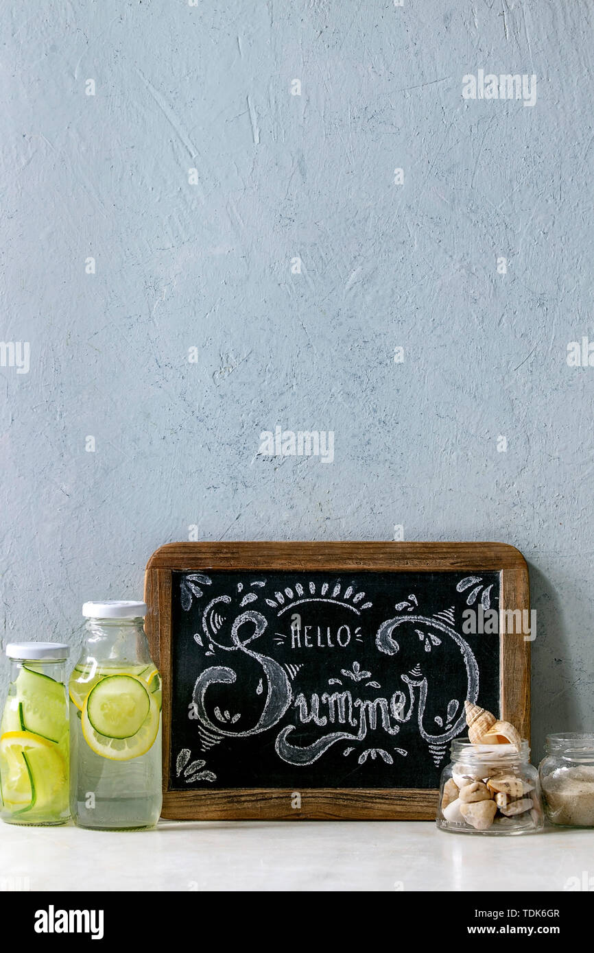 Summer theme. Chalk lettering Hello summer on vintage chalkboard, seashells and sand in glass jars, sunglasses, two glass bottles with sassy water on Stock Photo