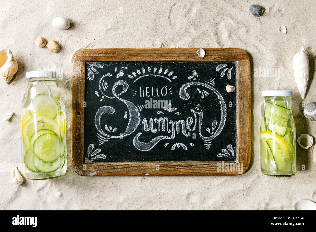 Summer theme. Chalk lettering Hello summer on vintage chalkboard, shells, sea stones, two glass bottles with sassy water on white sand as background. Stock Photo