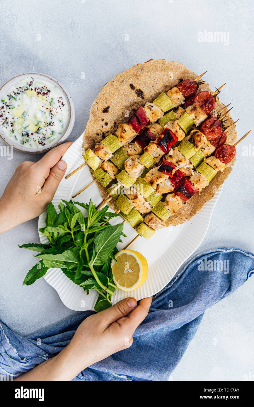 Woman serving spicy chicken kabobs on lavash bread accompanied by herbs, yogurt dip and lemon. Stock Photo