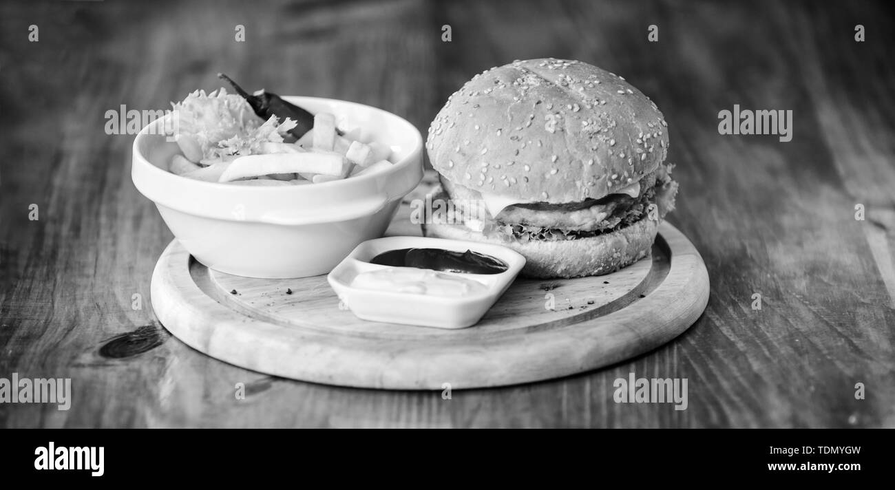 Cheat meal. Delicious burger with sesame seeds. Burger with cheese meat and salad. Fast food concept. Burger menu. High calorie snack. Hamburger and french fries and tomato sauce on wooden board. Stock Photo