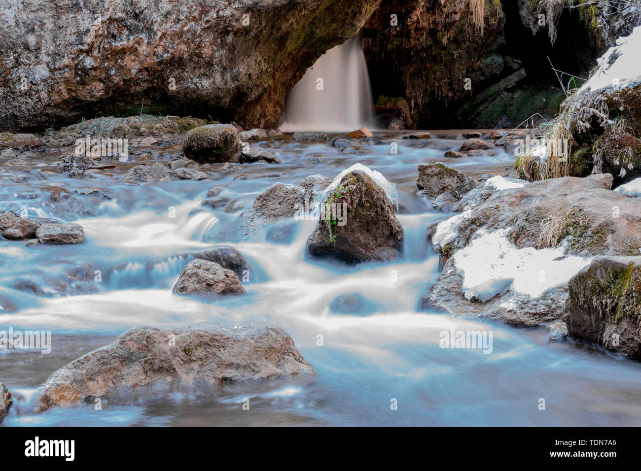 Cold mountain river flows between stones with snow and ice, selective focus, waterfall in the background, long exposure, Karachay-Cherkess Republic. Stock Photo