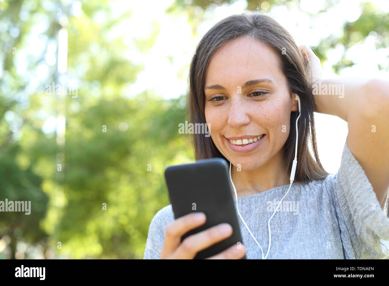 Happy adult woman wearing earphones listening to music checking smart phone standing in a park Stock Photo