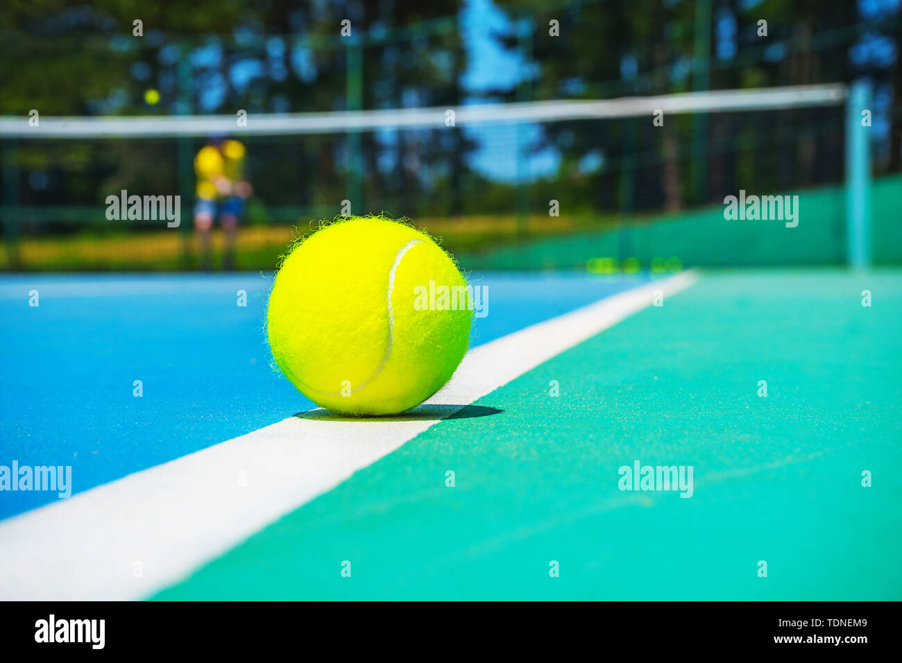 Tennis ball on white court line on hard modern blue green court with player, net, balls, trees on the background. Stock Photo
