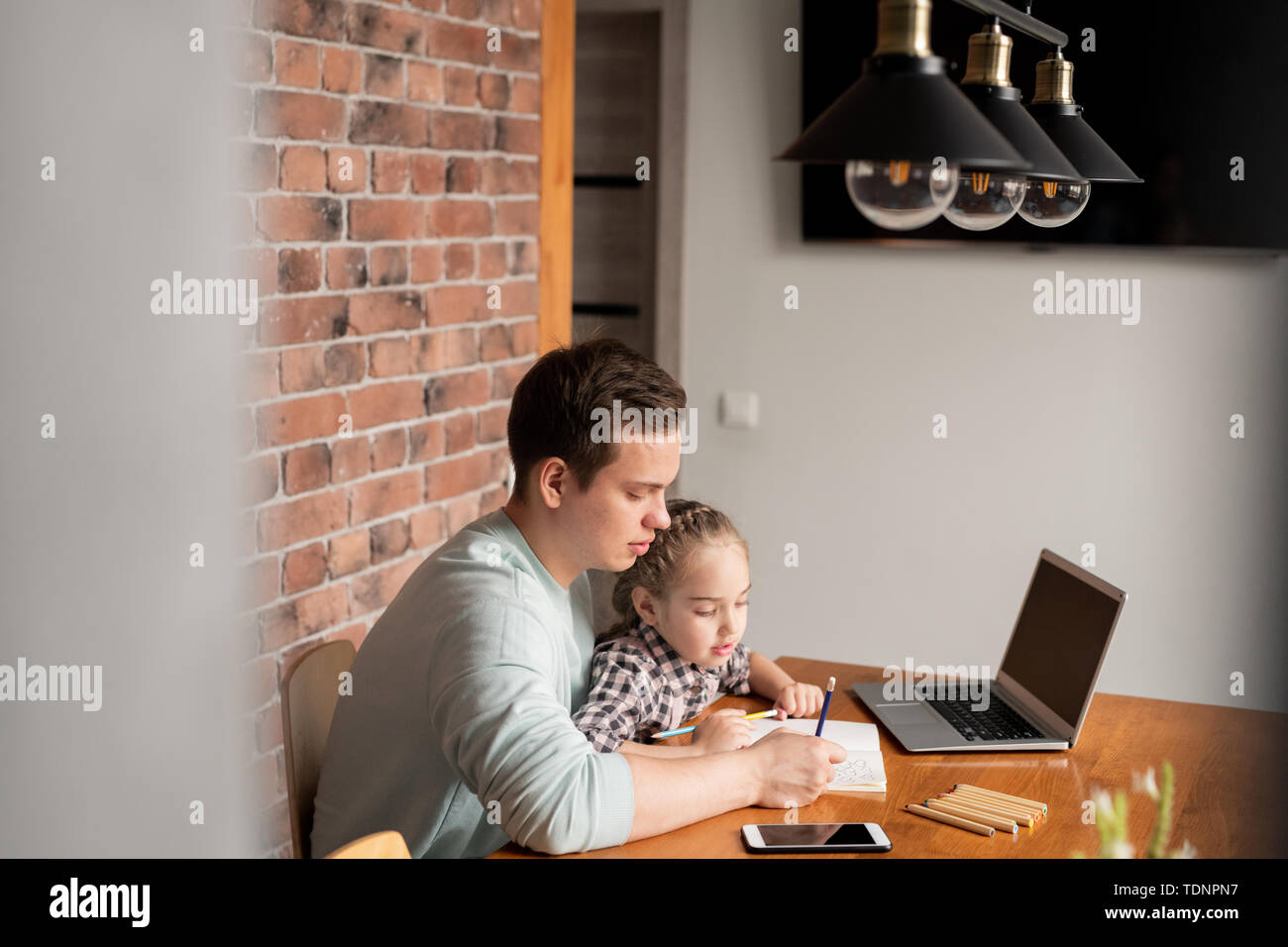 Serious concentrated young father sitting at table with devices and drawing hearts in notebook with daughter at home Stock Photo