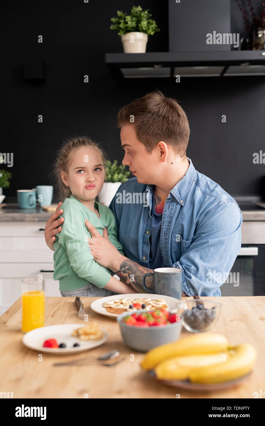 Picky daughter sitting on fathers knees and grimacing while refusing to eat breakfast made by father Stock Photo