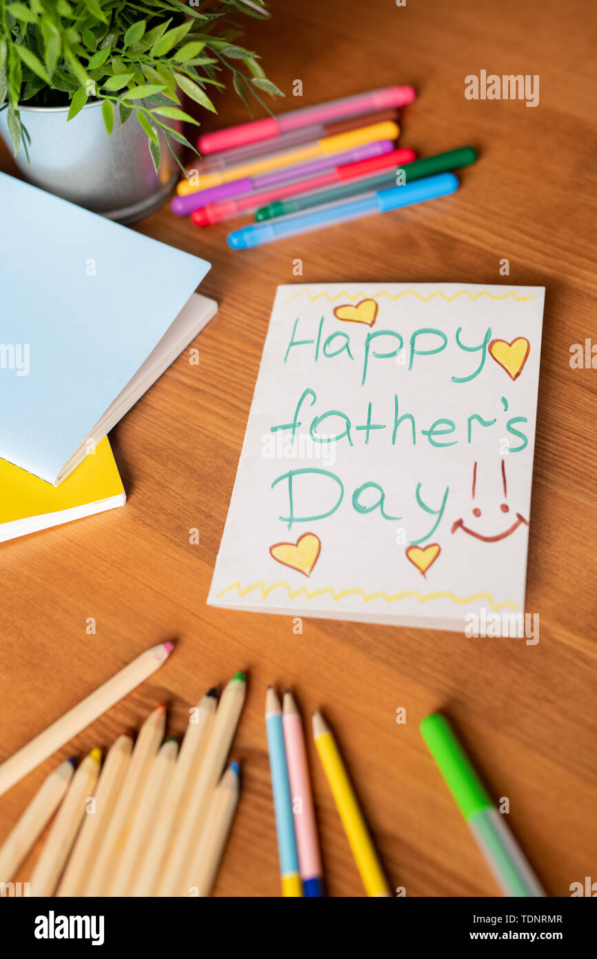 Close-up of handmade fathers day card with yellow hearts and colorful pencils and felt-tip pens on wooden table with potted plant and notebooks Stock Photo