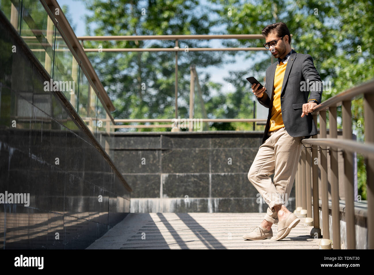 Young casual man in beige jeans, yellow pullover and dark blue jacket scrolling in smartphone by railings outdoors Stock Photo