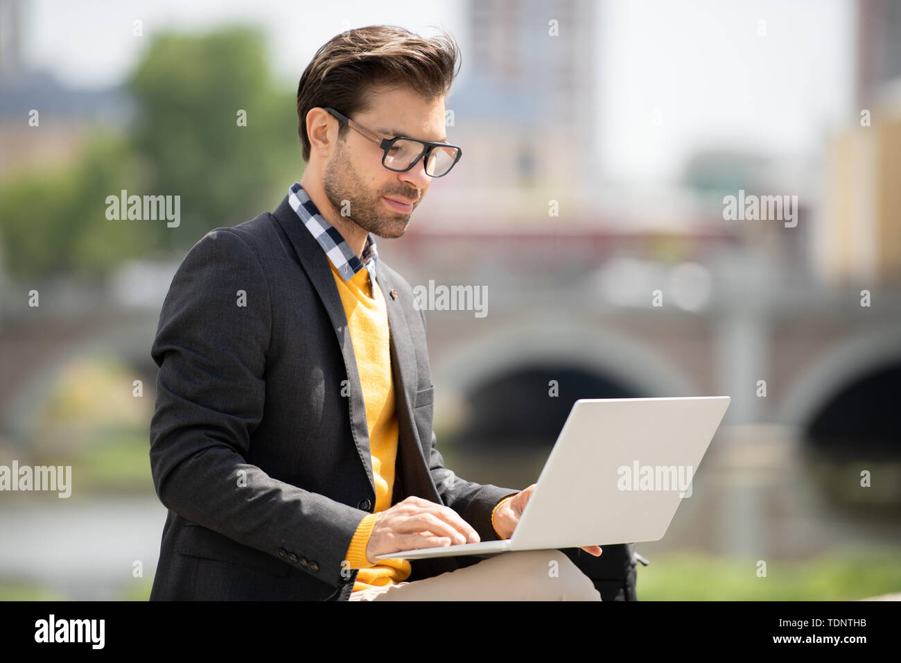 Handsome young freelancer in smart casualwear looking at laptop display while searching for online information outdoors Stock Photo