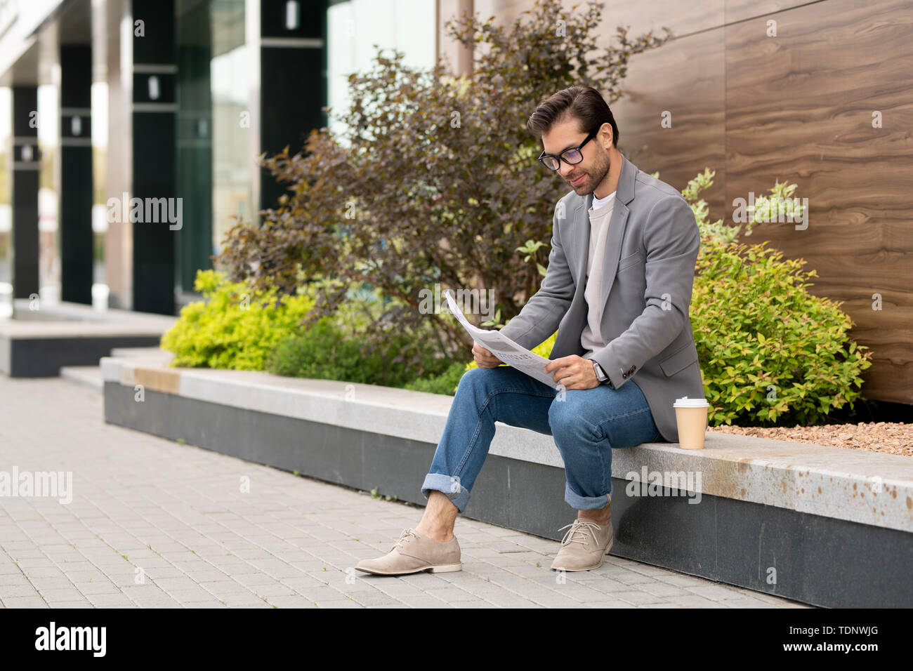 Young entrepreneur in smart casual sitting in urban environment and looking through newspaper with fresh news Stock Photo