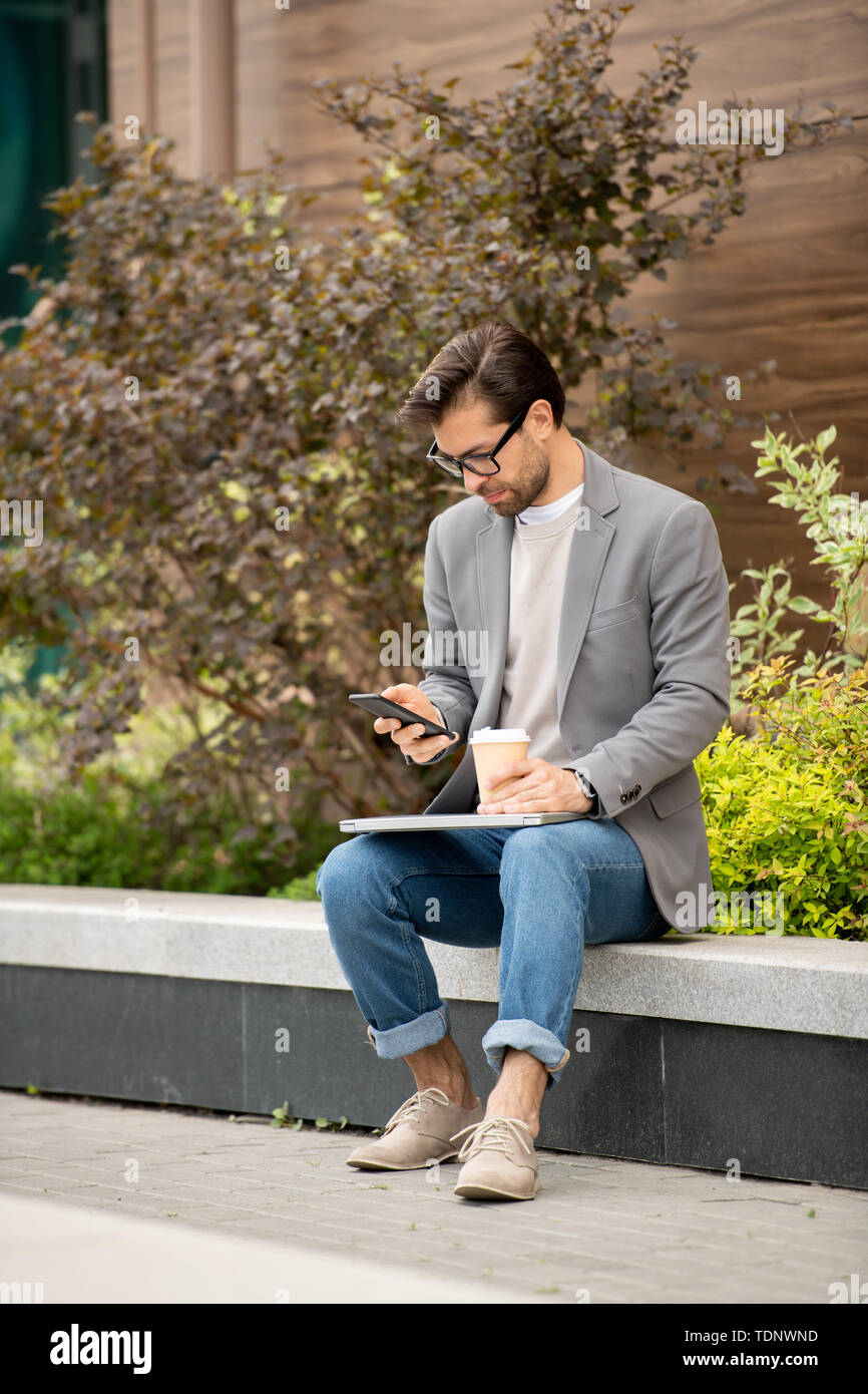 Busy young manager in smart casual using smartphone while having coffee outdoors in the morning Stock Photo