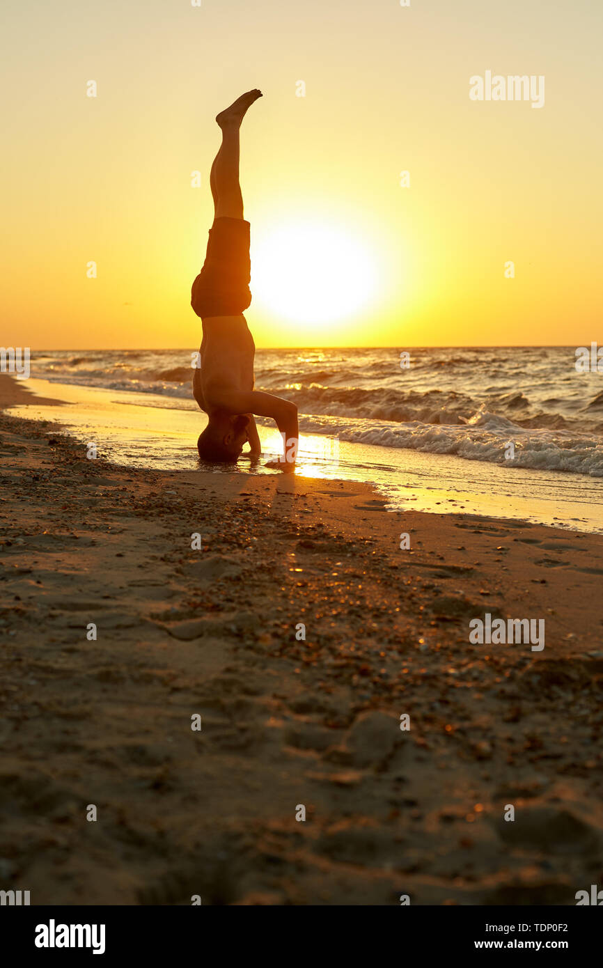 Young man practice balance asana on summer yoga session on beautiful golden beach of tropical island. Guy doing Salamba Sirsasana supported headstand Stock Photo