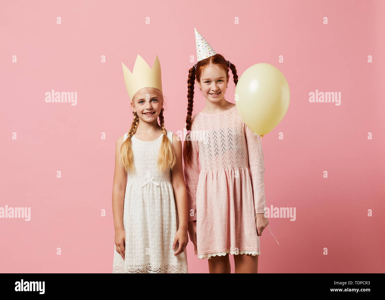 Portrait of two girls wearing party hats posing against pink background ...