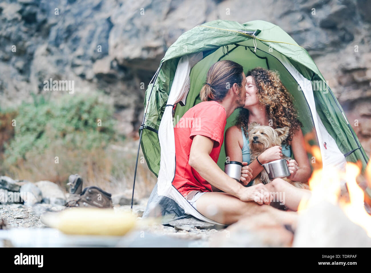 Travel couple camping in rock mountains after a trekking day - Happy people drinking hot tea inside tent with their dog next to bonfire Stock Photo