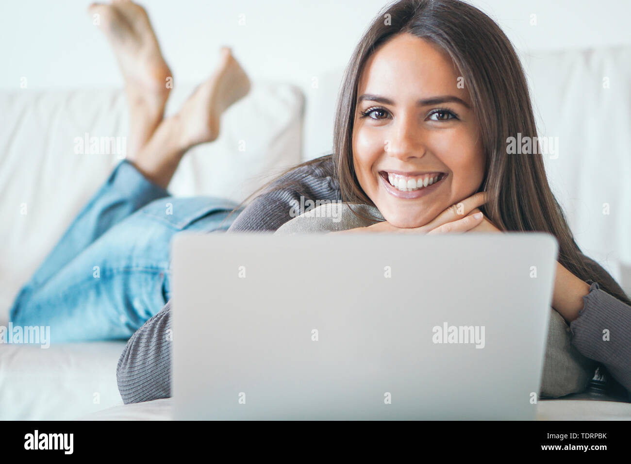 Young woman lying on sofa and using laptop at home - Happy girl surfing online with her computer while smiling at camera Stock Photo