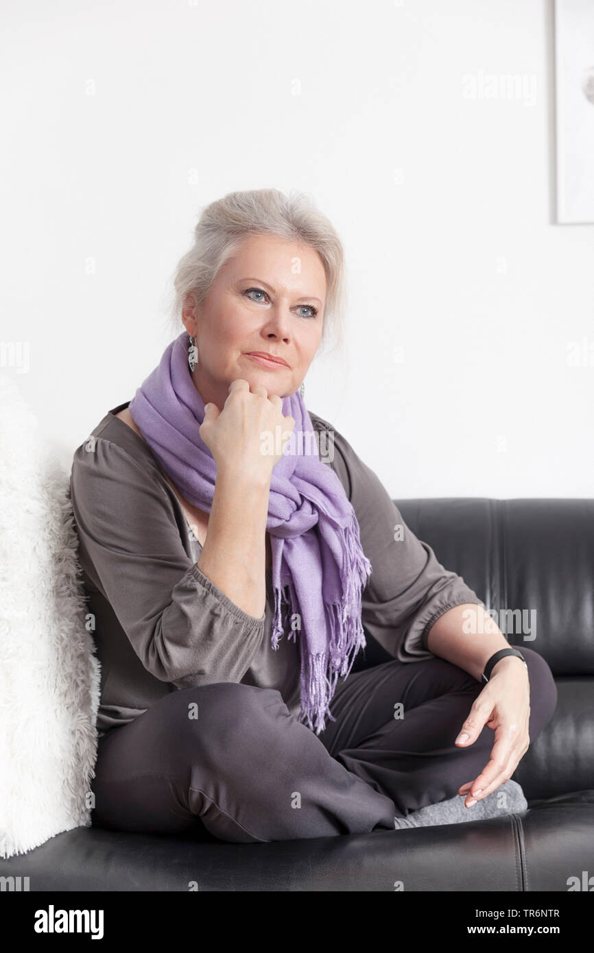 female senior sitting contemplatively in tailor seat on a sofa, Germany Stock Photo
