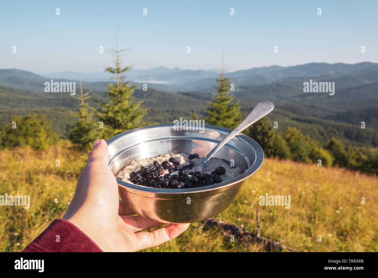 Breakfast in the hike, a bowl of porridge and berries in the female hand on the background of the morning mountain landscape Stock Photo