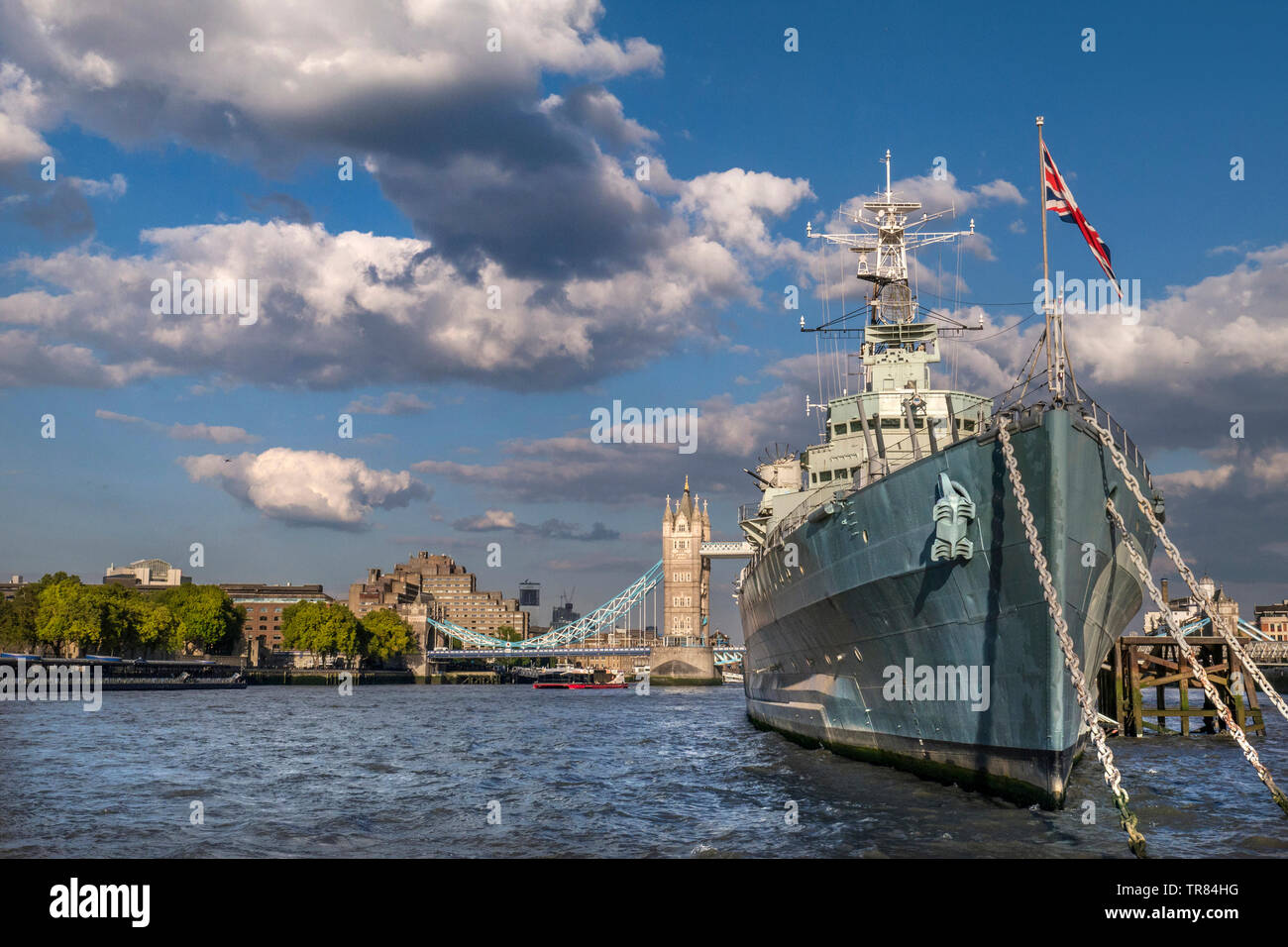 HMS Belfast ship flying Union Jack Flag, moored on River Thames in late afternoon sunlight, with Tower Bridge and The Tower Hotel behind London SE1 Stock Photo