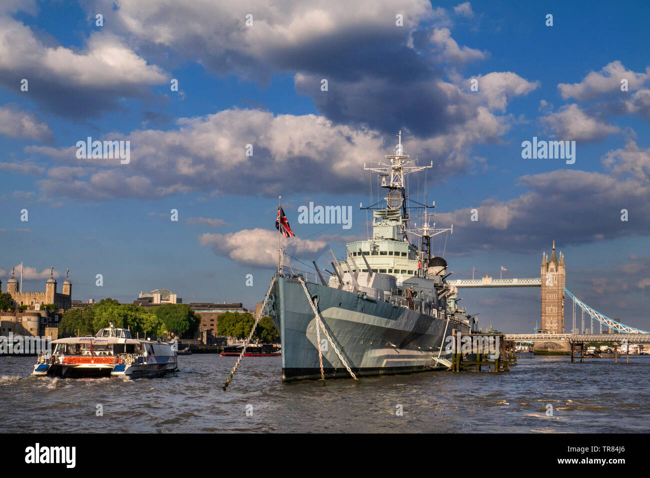 HMS Belfast museum tourist ship moored on River Thames with The Tower of London, a passing RB1 River Clipper Boat and Tower Bridge behind London SE1 Stock Photo