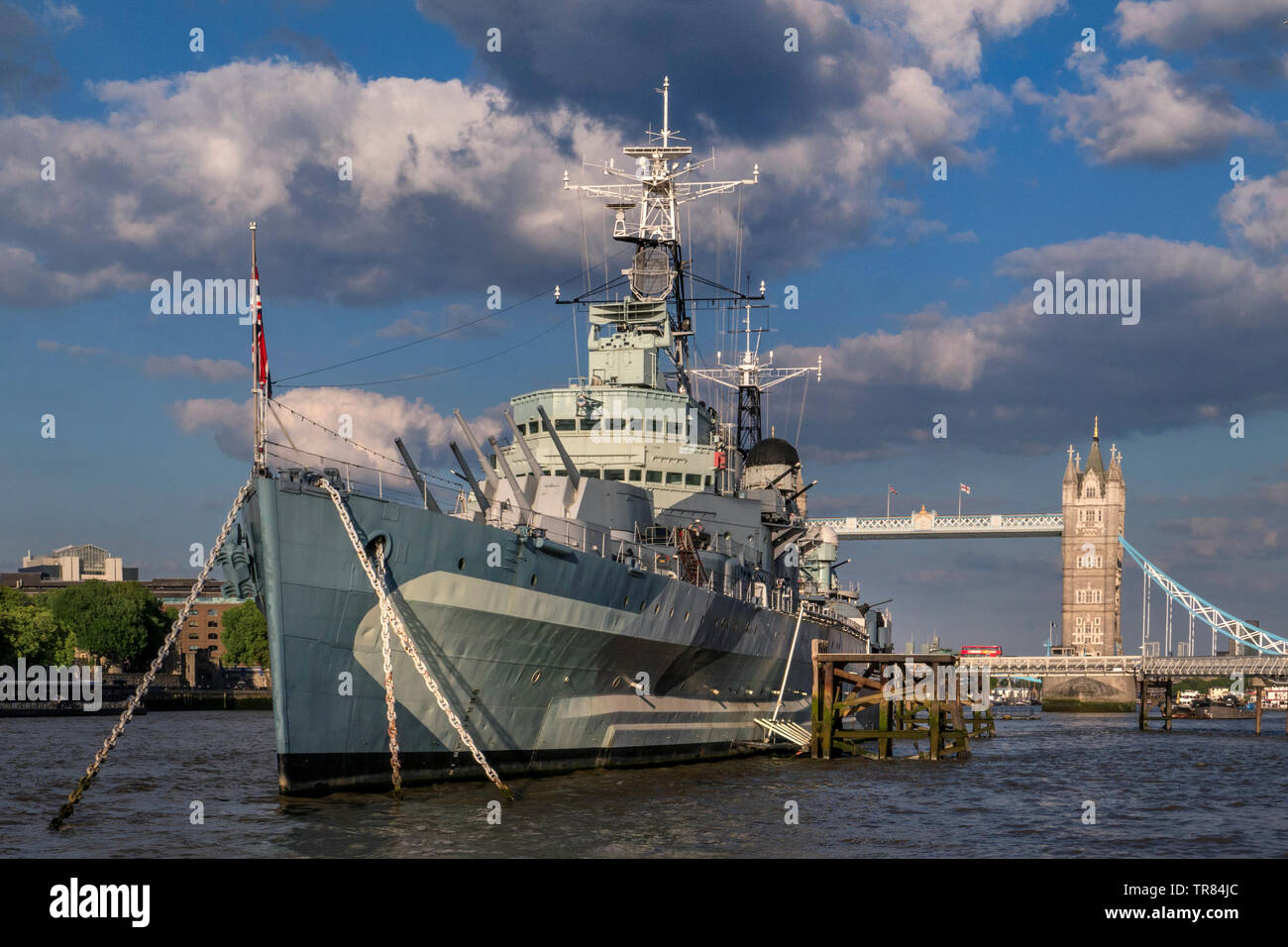 HMS Belfast tourist attraction ship, moored on River Thames in late afternoon sunlight, with Tower Bridge and London Red Bus crossing London SE1 Stock Photo
