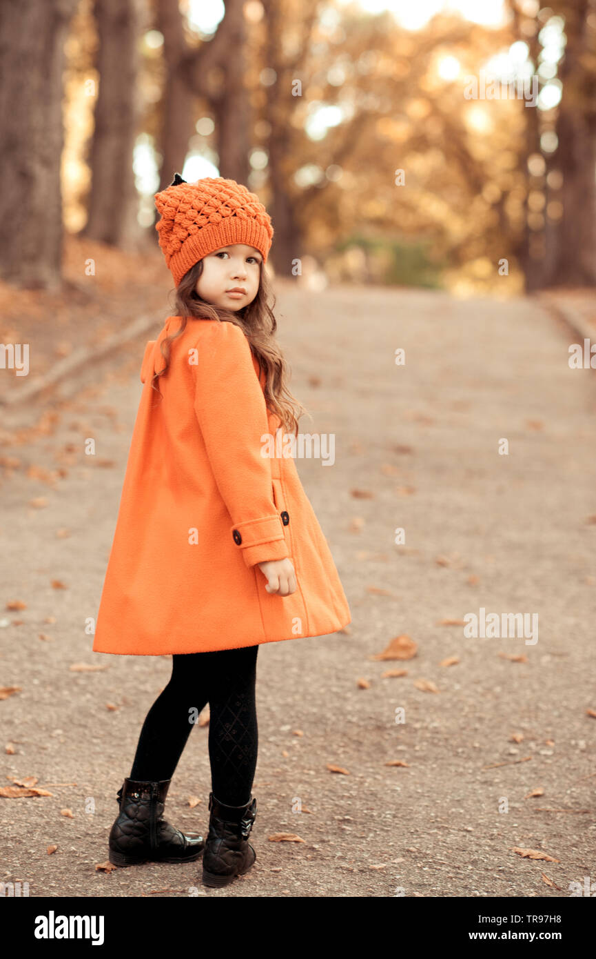 Kid girl 4-5 year old wearing stylish coat and knitted hat in park. Standing outdoors. Looking at camera. Childhood. Stock Photo