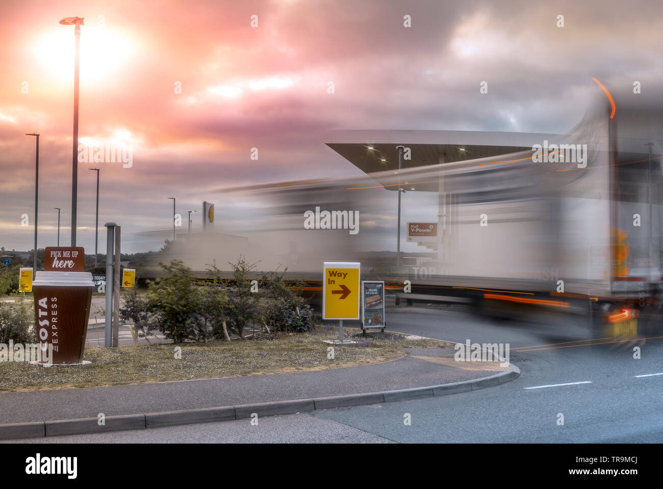 A large Articulated Lorry pulls onto a garage forecourt, as the sun illuminates storm clouds gathering in the sky above. Stock Photo