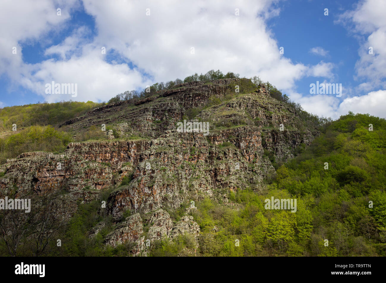 Impressive, red, rocky peak on Old mountain (Stara planina) in Serbia, covered by vivid green, sunlit trees and bushes during early spring Stock Photo