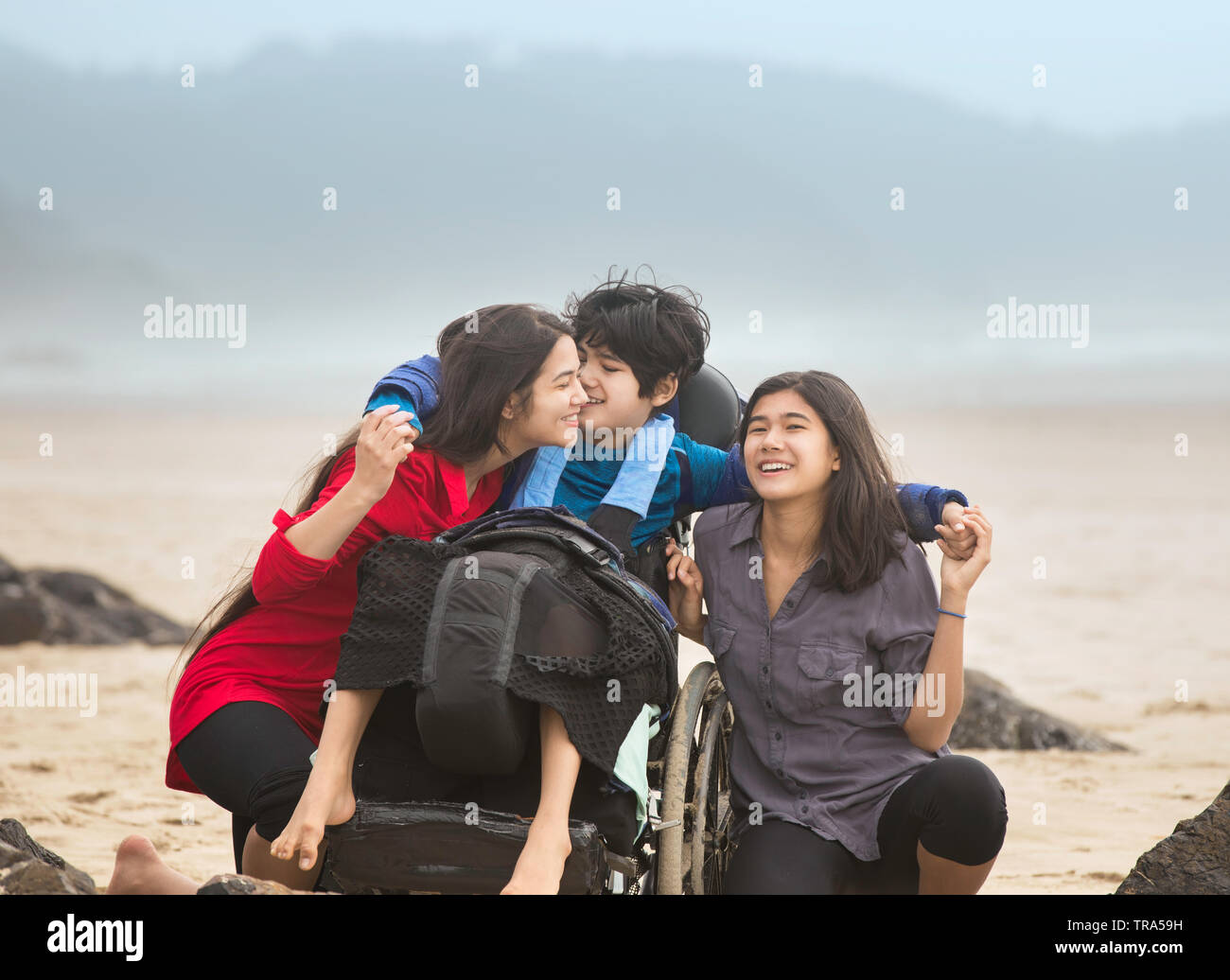 Disabled younger brother in wheelchair laughing and giving older sistesr a hug while they sit together by ocean on misty foggy beach Stock Photo