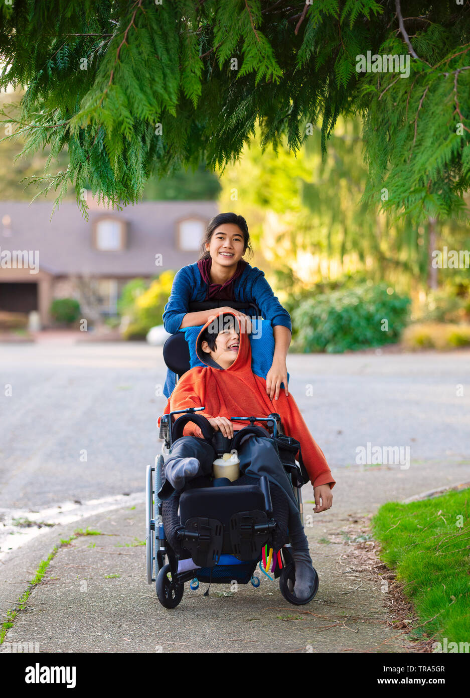 Big sister pushing disabled little brother in wheelchair around neighborhood, laughing and smiling Stock Photo