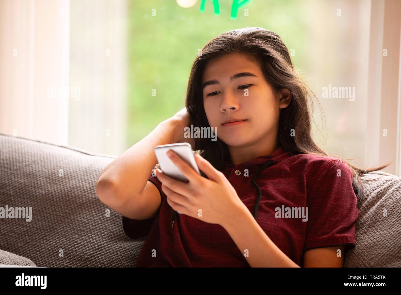 Biracial teen girl sitting on gray couch by large picture window, looking at smartphone Stock Photo