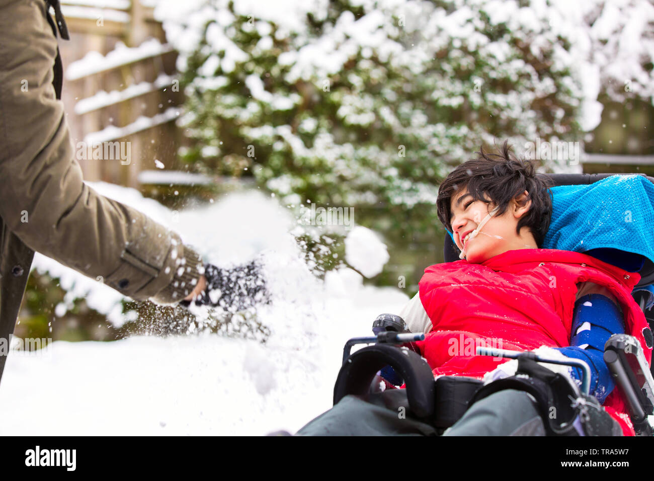 Cute biracial disabled twelve year old boy in wheelchair sitting outside in winter, playing with an adult throwing snow and smiling Stock Photo